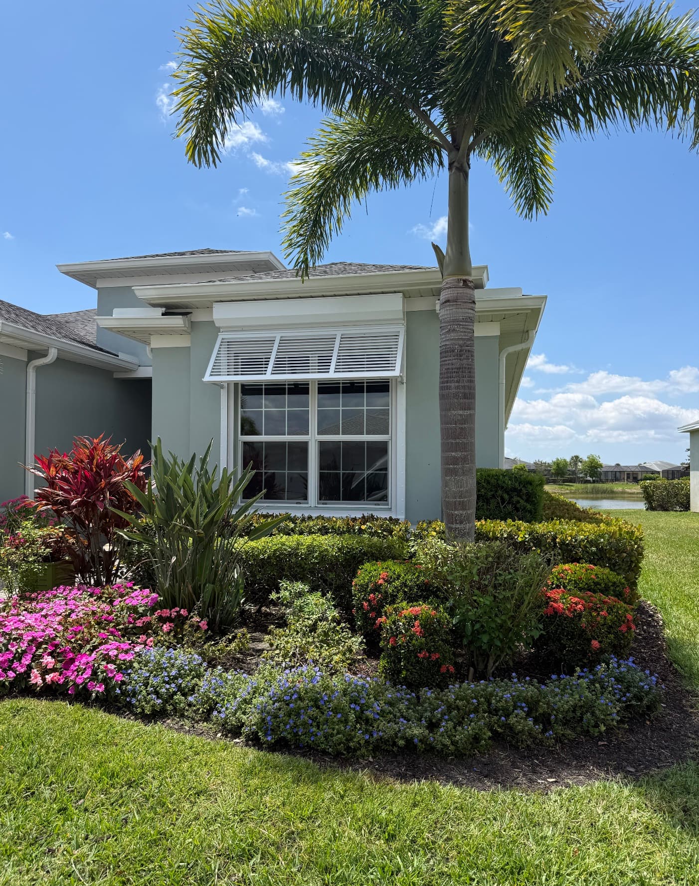 White Bahama shutter propped open over a double window on a light blue-gray home, with a palm tree and flower beds in front