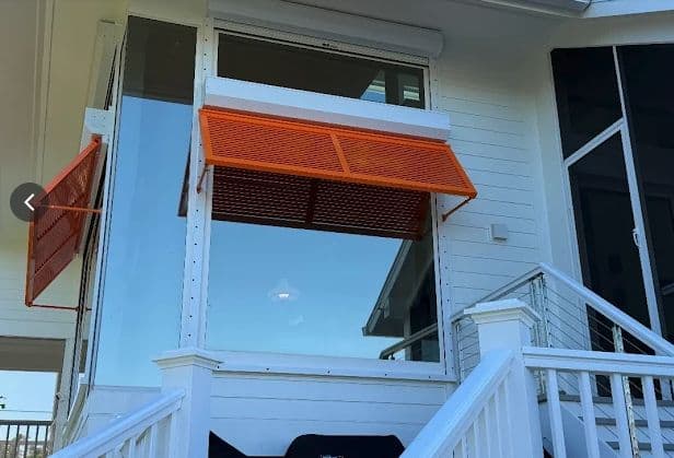 Orange Bahama shutter propped open over a window on a white raised home