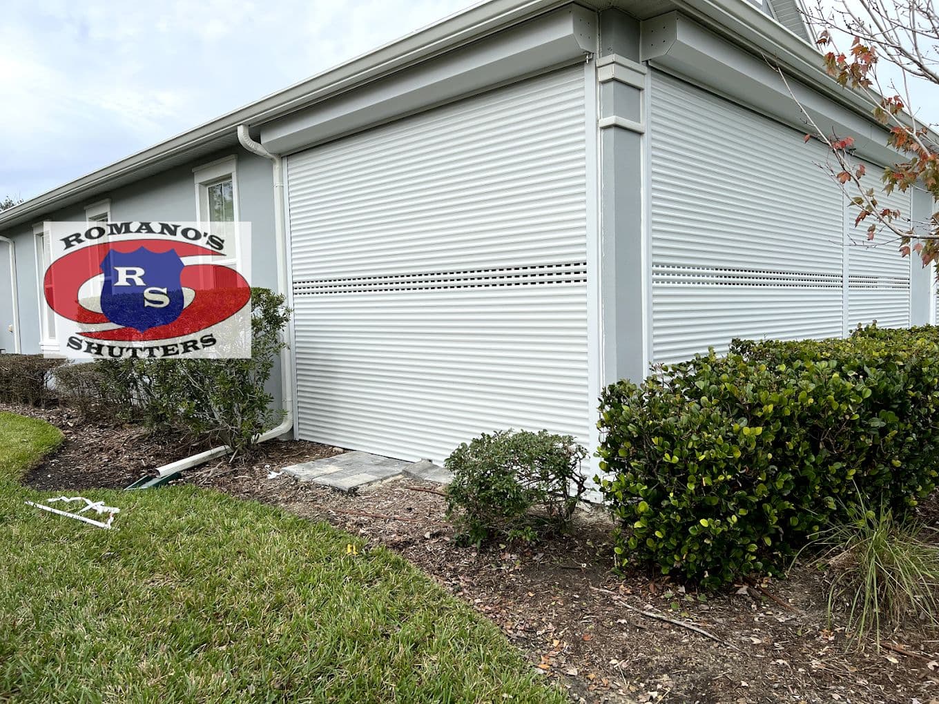Closed white roll-down hurricane shutters covering the side of a home, with a Romano's Shutters yard sign
